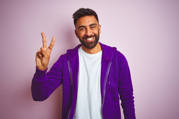 Young indian man wearing purple sweatshirt standing over isolated pink background showing and pointing up with fingers number two while smiling confident and happy.