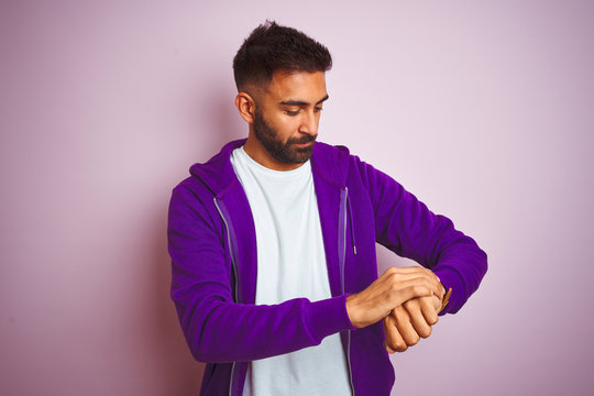 Young indian man wearing purple sweatshirt standing over isolated pink background Checking the time on wrist watch, relaxed and confident