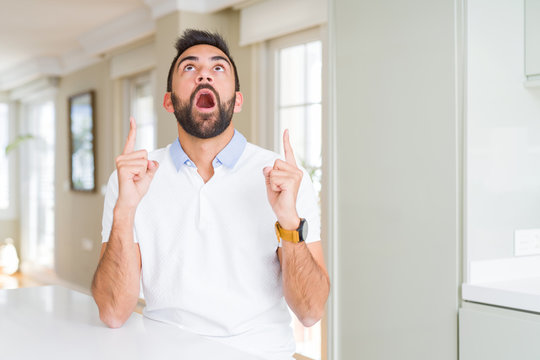 Handsome hispanic man casual white t-shirt at home amazed and surprised looking up and pointing with fingers and raised arms.