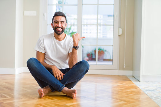 Handsome hispanic man wearing casual t-shirt sitting on the floor at home smiling with happy face looking and pointing to the side with thumb up.