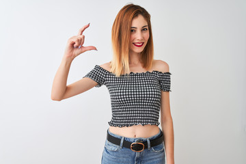 Beautiful redhead woman wearing casual t-shirt standing over isolated white background smiling and confident gesturing with hand doing small size sign with fingers looking and the camera.