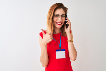 Redhead businesswoman wearing id card talking on smartphone over isolated white background happy with big smile doing ok sign, thumb up with fingers, excellent sign