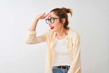 Redhead woman wearing striped shirt and glasses standing over isolated white background very happy and smiling looking far away with hand over head. Searching concept.