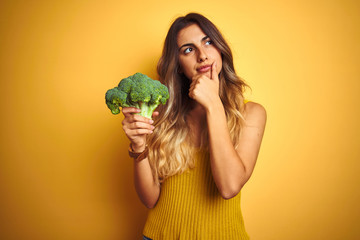 Young beautiful woman eating broccoli over yellow isolated background serious face thinking about...