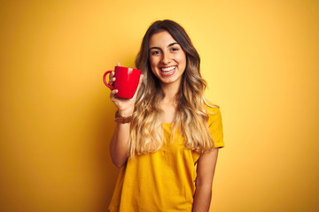 Young beautiful woman holding  red cup of coffee over yellow isolated background with a happy face standing and smiling with a confident smile showing teeth
