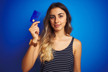 Young beautiful woman holding credit card over blue isolated background with a confident expression on smart face thinking serious