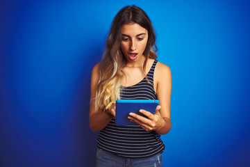 Young beautiful woman using touchpad tablet over blue isolated background scared in shock with a surprise face, afraid and excited with fear expression