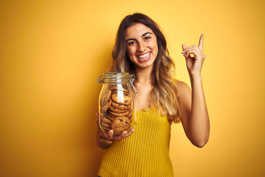 Young beautiful woman holding jar of cookies over yellow isolated background very happy pointing with hand and finger to the side