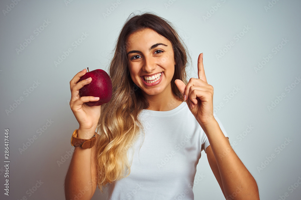 Wall mural young beautiful woman eating red apple over grey isolated background surprised with an idea or quest