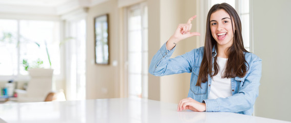 Wide angle picture of beautiful young woman sitting on white table at home smiling and confident gesturing with hand doing size sign with fingers while looking and the camera. Measure concept.