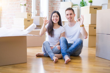 Young beautiful couple sitting on the floor at new home around cardboard boxes doing ok sign with fingers, excellent symbol