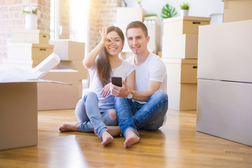 Fototapeta na wymiar Beautiful couple sitting on the floor using smartphone at new home around cardboard boxes with happy face smiling doing ok sign with hand on eye looking through fingers