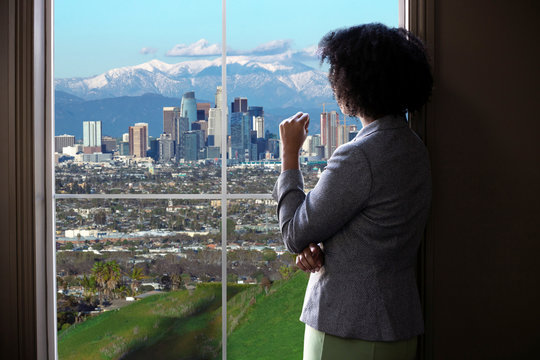 Black female business woman looking out the window of an office in Los Angeles.  She looks like a female architect thinking of urban development or a city mayor or governor planning zoning laws.