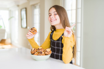 Beautiful young girl eating chocolate chips muffins very happy pointing with hand and finger to the...