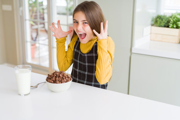 Beautiful young girl kid eating chocolate cereals and glass of milk for breakfast celebrating mad...