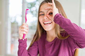 Beautiful young girl kid holding pink dental toothbrush with happy face smiling doing ok sign with hand on eye looking through fingers