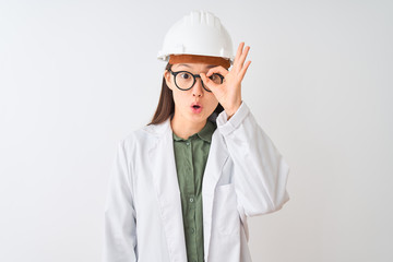 Young chinese engineer woman wearing coat helmet glasses over isolated white background doing ok gesture shocked with surprised face, eye looking through fingers. Unbelieving expression.