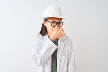 Young chinese engineer woman wearing coat helmet glasses over isolated white background smelling...