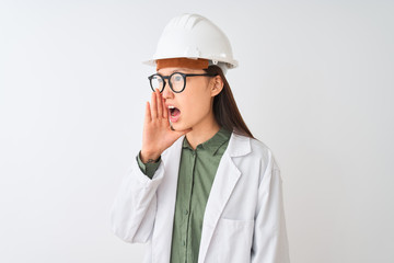 Young chinese engineer woman wearing coat helmet glasses over isolated white background shouting and screaming loud to side with hand on mouth. Communication concept.