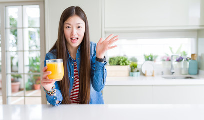 Beautiful Asian woman drinking a glass of fresh orange juice very happy and excited, winner expression celebrating victory screaming with big smile and raised hands