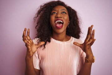 Young african american woman wearing t-shirt standing over isolated pink background crazy and mad shouting and yelling with aggressive expression and arms raised. Frustration concept.