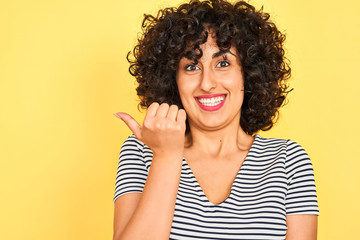 Young arab woman with curly hair wearing striped dress over isolated yellow background pointing and showing with thumb up to the side with happy face smiling