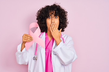 Young arab doctor woman with curly hair holding cancer ribbon over isolated pink background cover mouth with hand shocked with shame for mistake, expression of fear, scared in silence, secret concept