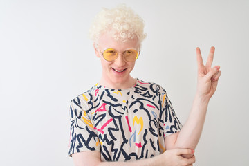 Young albino blond man wearing colorful t-shirt and sunglasses over isolated red background smiling with happy face winking at the camera doing victory sign. Number two.