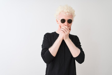 Young albino man wearing black t-shirt and sunglasess standing over isolated white background shocked covering mouth with hands for mistake. Secret concept.