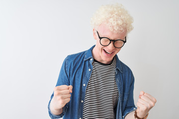 Young albino blond man wearing denim shirt and glasses over isolated white background very happy and excited doing winner gesture with arms raised, smiling and screaming for success. 