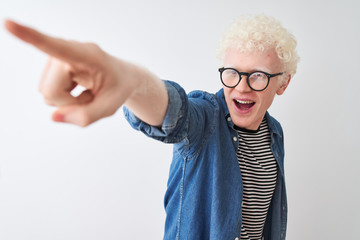 Young albino blond man wearing denim shirt and glasses over isolated white background Pointing with finger surprised ahead, open mouth amazed expression, something on the front