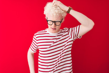 Young albino blond man wearing striped t-shirt and glasses over isolated red background confuse and wonder about question. Uncertain with doubt, thinking with hand on head. Pensive concept.