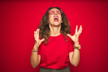 Middle age senior woman wearing cute heart shaped glasses over red isolated background crazy and...