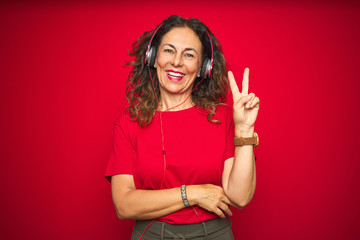 Middle age senior woman wearing headphones listening to music over red isolated background smiling...