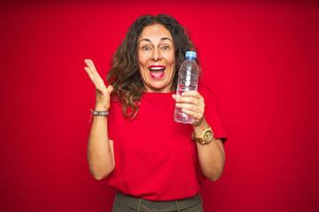 Middle age senior woman holding plastic water bottle over red isolated background very happy and excited, winner expression celebrating victory screaming with big smile and raised hands