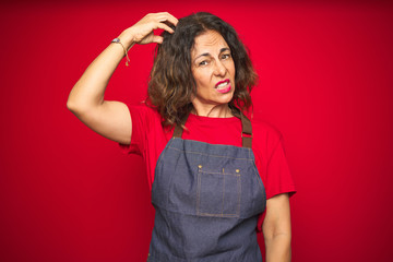 Middle age senior woman wearing apron uniform over red isolated background confuse and wondering about question. Uncertain with doubt, thinking with hand on head. Pensive concept.