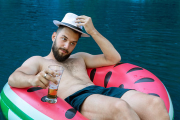 young attractive man takes off his hat, welcomes. Swims lying in an inflatable circle with a glass of beer