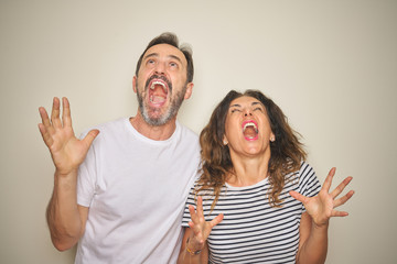 Beautiful middle age couple together standing over isolated white background crazy and mad shouting and yelling with aggressive expression and arms raised. Frustration concept.