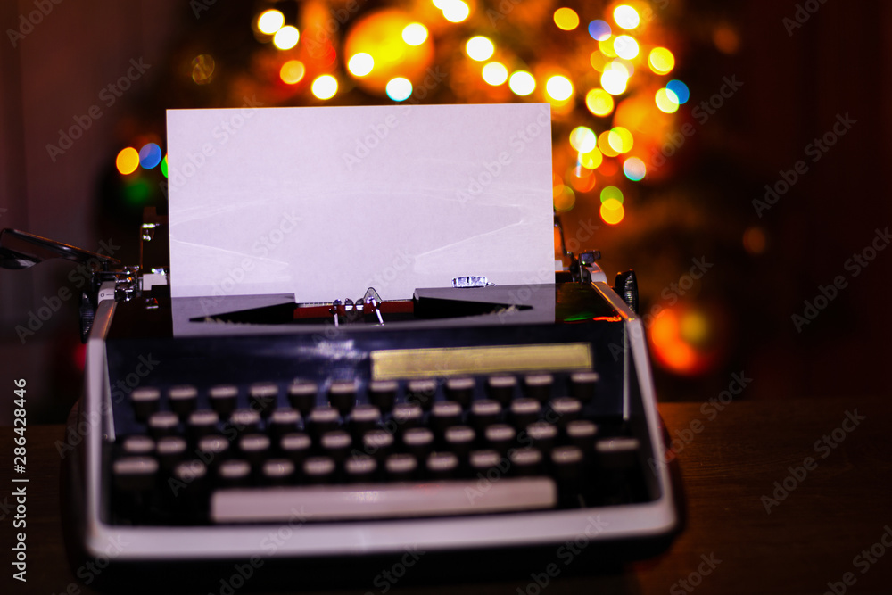 Fototapete Retro typewriter with a blank sheet of paper for text. Dark Christmas and New Year background. Mock up, Copy space with a glowing bokeh garland in the background.