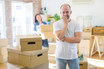 Young couple arround cardboard boxes moving to a new house, bald man standing at home looking stressed and nervous with hands on mouth biting nails. Anxiety problem.