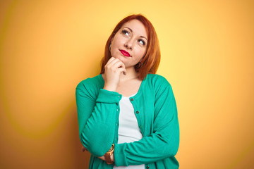 Youg beautiful redhead woman wearing winter green sweater over isolated yellow background with hand on chin thinking about question, pensive expression. Smiling with thoughtful face. Doubt concept.