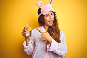 Young woman wearing pajama and sleep mask drinking water over yellow isolated background very happy pointing with hand and finger