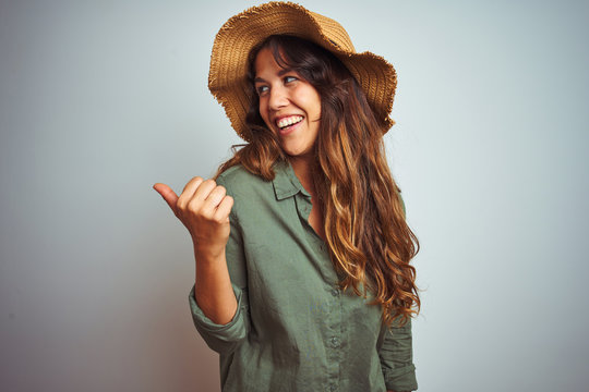 Young beautiful woman on vacation wearing green shirt and hat over white isolated background smiling with happy face looking and pointing to the side with thumb up.