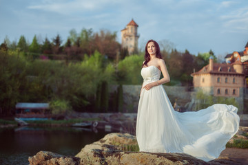 Fototapeta premium Beautiful young woman in a white dress stands on the banks of the river.