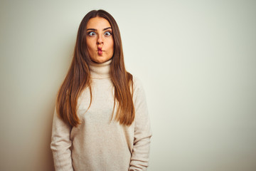 Obraz premium Young beautiful woman wearing winter turtleneck sweater over isolated white background making fish face with lips, crazy and comical gesture. Funny expression.