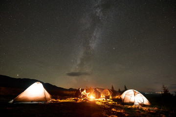 Three tourist tents on mountain valley and group of four people, young men and woman with guitar by burning bonfire on background of dark sky with bright sparkling stars and Milky Way constellation.