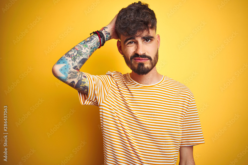 Poster Young man with tattoo wearing striped t-shirt standing over isolated yellow background confuse and wondering about question. Uncertain with doubt, thinking with hand on head. Pensive concept.