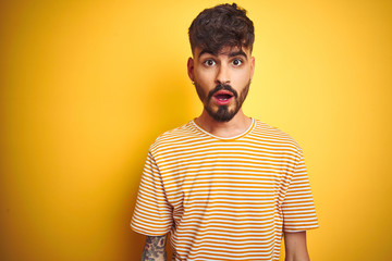 Young man with tattoo wearing striped t-shirt standing over isolated yellow background afraid and shocked with surprise and amazed expression, fear and excited face.