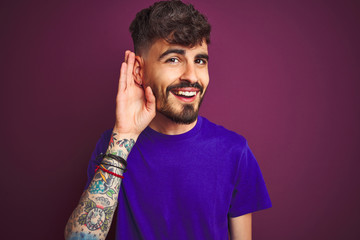 Young man with tattoo wearing t-shirt standing over isolated purple background smiling with hand over ear listening an hearing to rumor or gossip. Deafness concept.