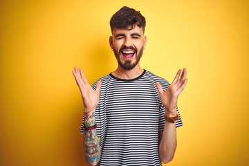 Young man with tattoo wearing striped t-shirt standing over isolated yellow background celebrating...